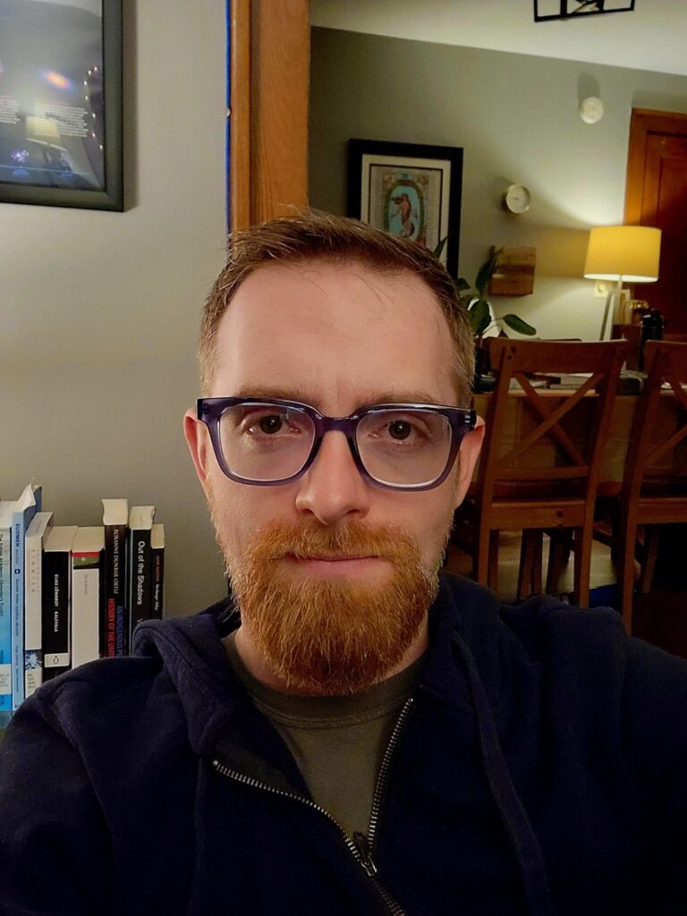 A second photograph of David. This photo is in his home. Several books can be seen over his right shoulder, and his dining room can be seen over his left. He has a short beard that was recently trimmed.