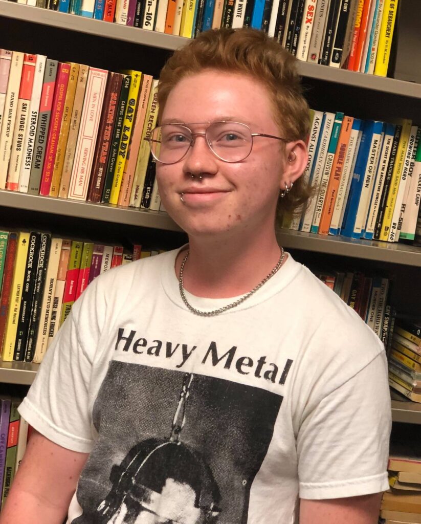 Photograph of Jimmy Arnold in front of Quatrefoil's collection of gay pulp novels. Jimmy wears glasses, has several piercings, and a chain around their neck. They are wearing a 'Heavy Metal' t-shirt.