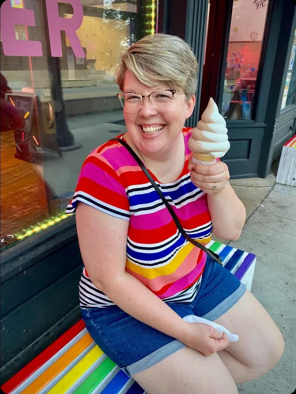 Katie Hagen in a multi-colored sweater eating a soft serve ice cream cone and sitting on a rainbow bench